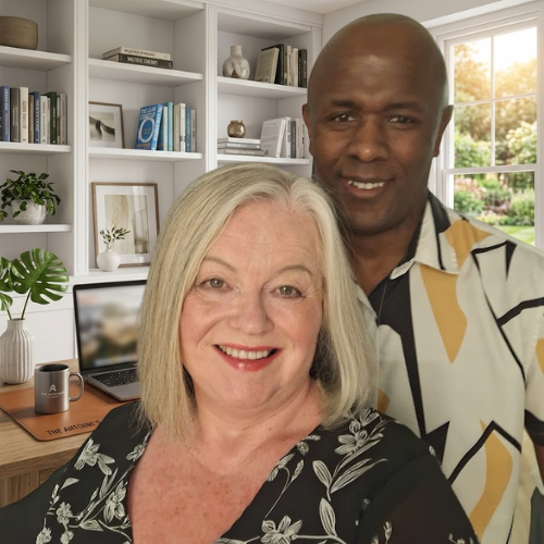 A smiling mature woman with shoulder-length blonde hair and a floral top, standing in front of a smiling Black man in a patterned shirt. They are positioned in a bright, modern home office setting with a bookshelf, a desk featuring a laptop and a mug with branding, and a window showing greenery in the background.